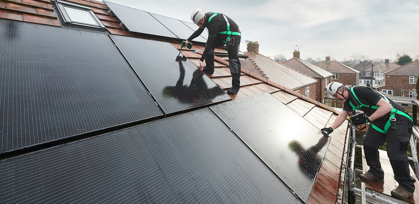 Two installers in hard hats and safety harnesses fitting black solar panels on a tiled roof, with brick houses in the background.
